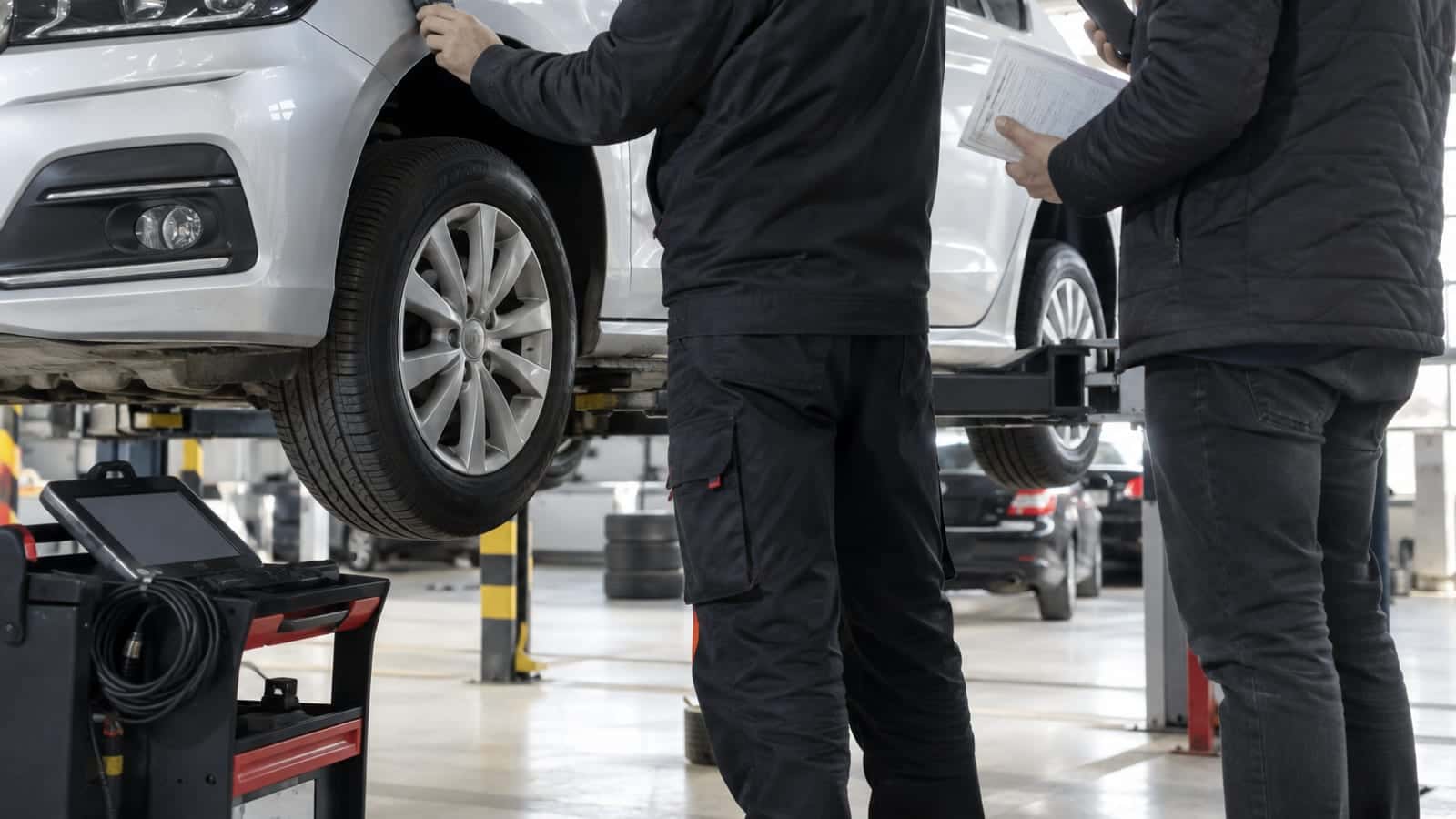 Ekspertiz atölyesinde lifte kaldırılmış otomobilin çamurluğuna boya ölçüm cihazı tutan teknisyen ve elinde ruhsatla bekleyen alıcı.
