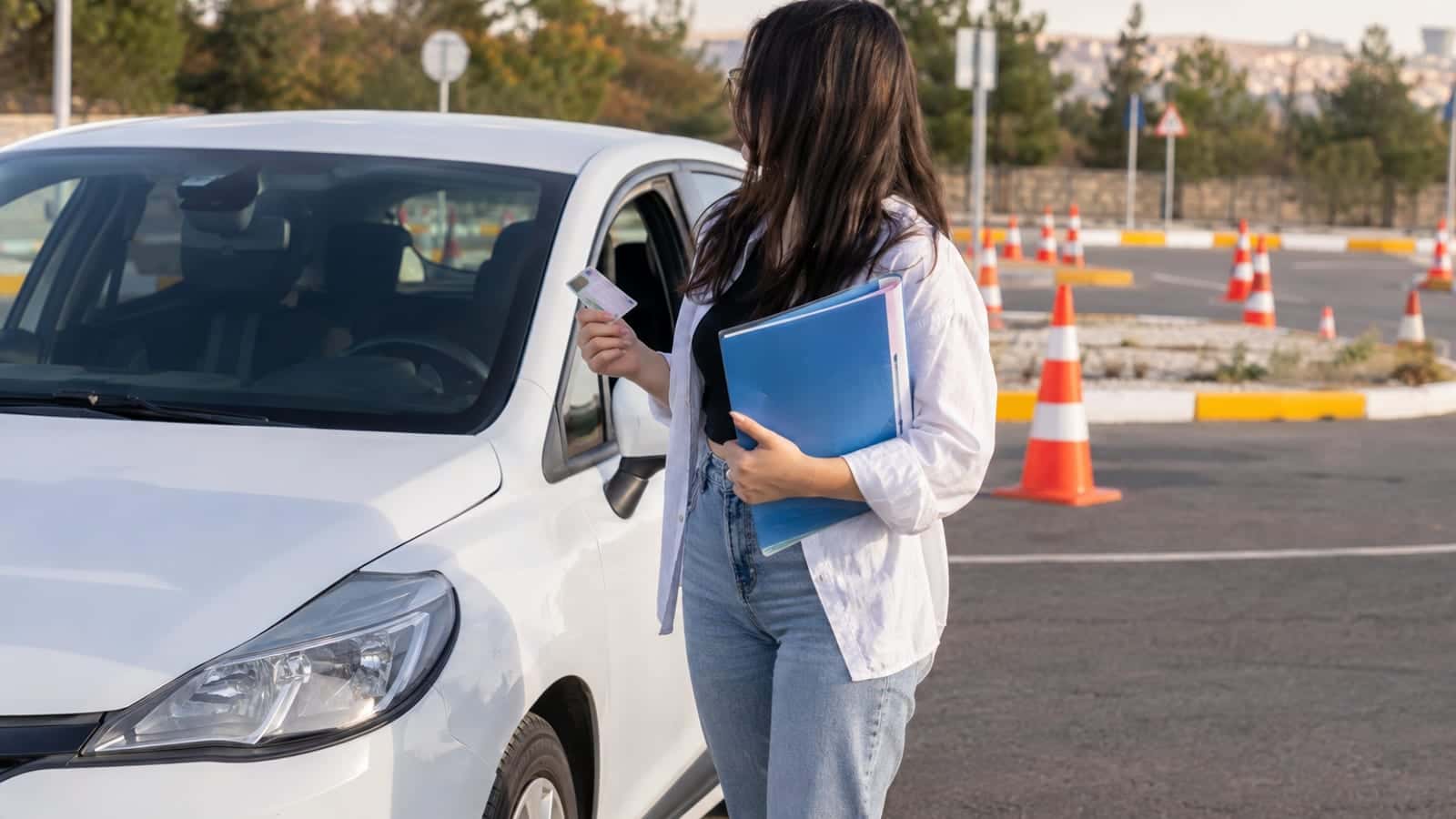 Sürücü kursu otoparkında eğitim otomobilinin yanında belge dosyası tutan genç bir kadın, arkada eğitim pisti konileri.