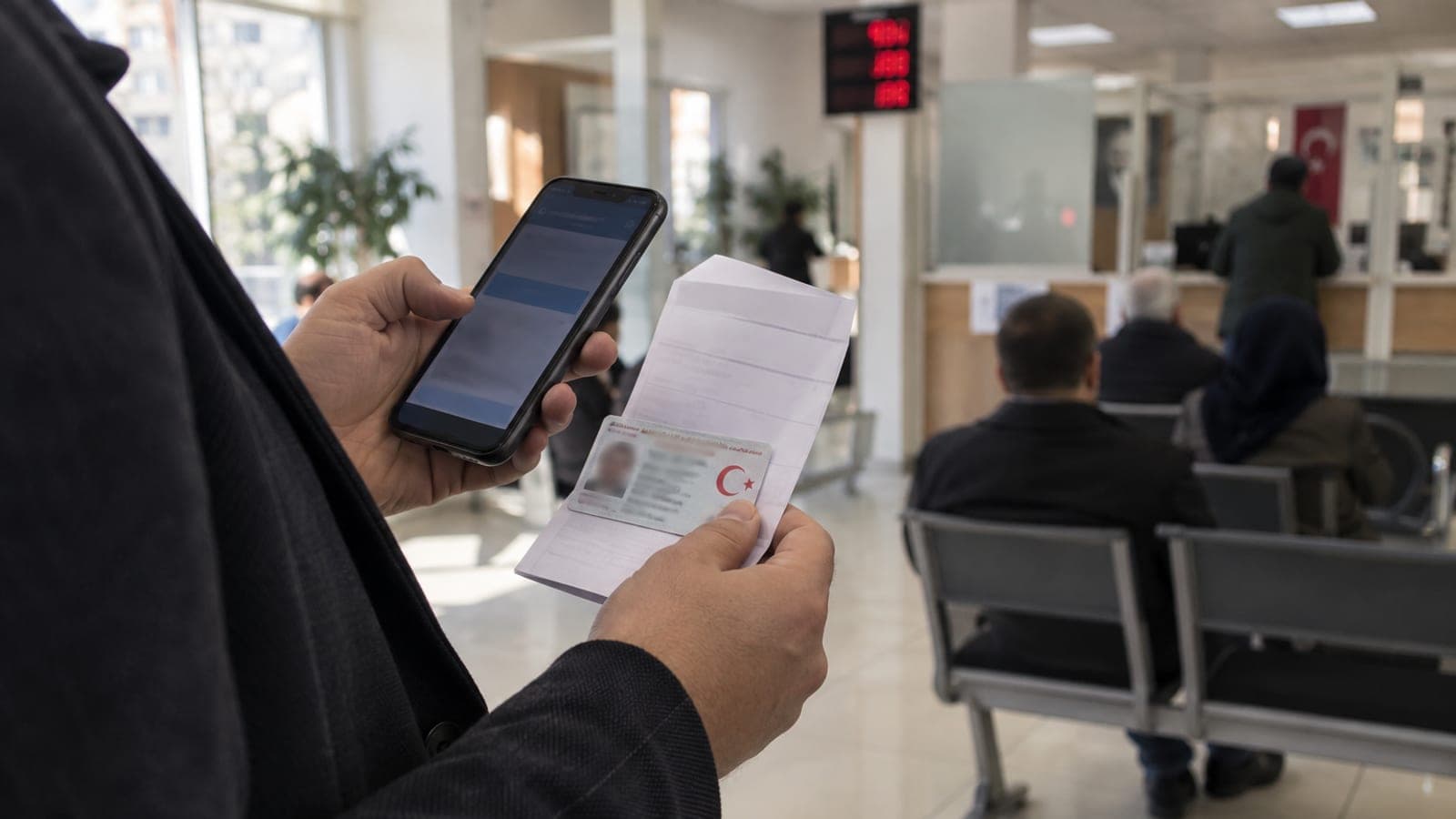 Kimlik kartı ve ödeme dekontu tutan kişi, telefonundaki bankacılık uygulamasına bakarken kurum koridorunda bekliyor