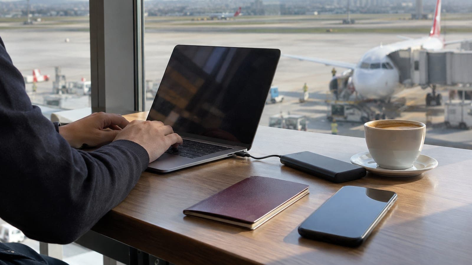 Havalimanı bekleme salonunda masada açık duran ince bir dizüstü bilgisayar, yanında pasaport, kahve ve powerbank