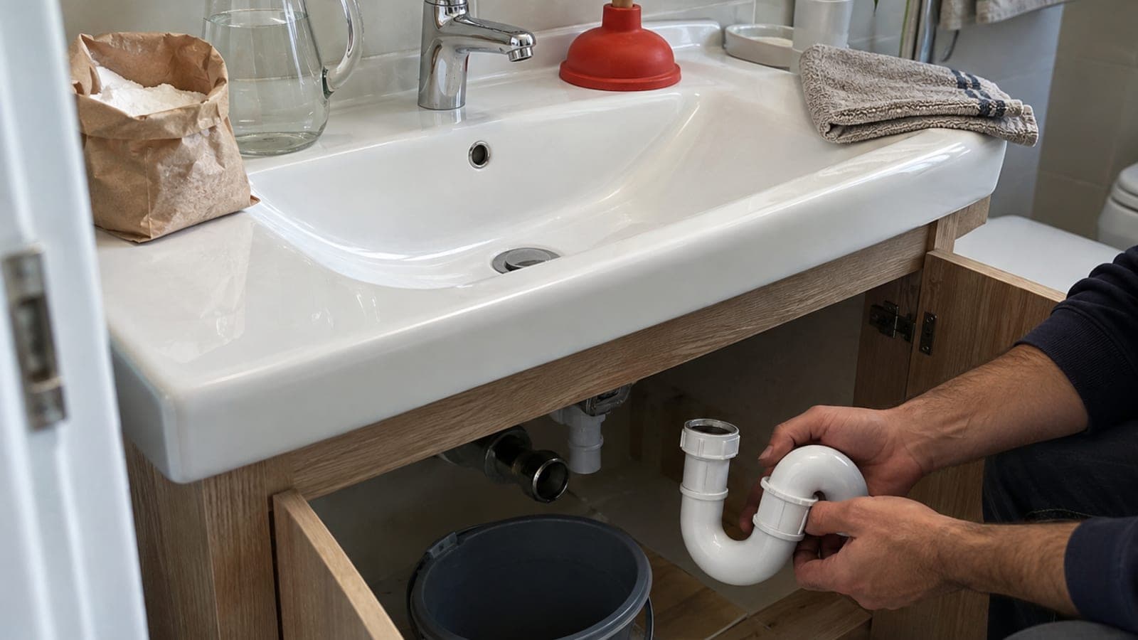Banyo lavabosu kenarında karbonat paketi, sirke sürahisi ve kırmızı lastik pompa; altta kova ile sökülmüş U dirsek sifon.