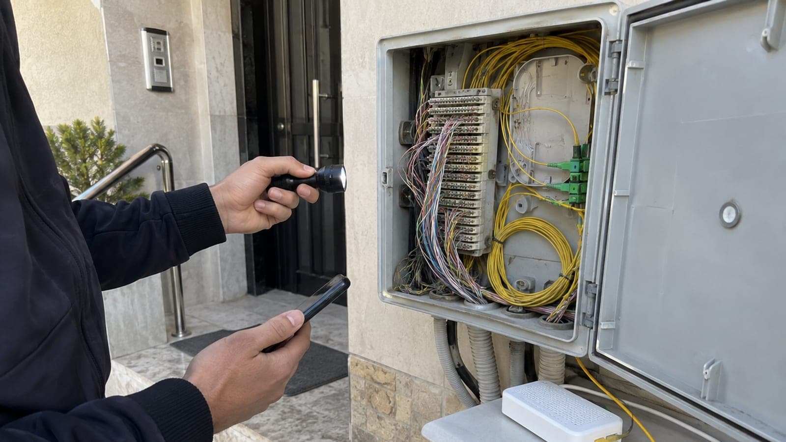 Bir kişi apartman girişindeki dağıtım kutusunu el feneriyle inceliyor; kutuda bakır telefon telleri ve sarı fiber kablolar görünüyor
