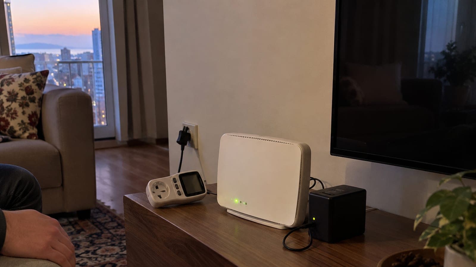Karanlık salonda TV sehpası üstünde çalışan beyaz modem, yanında mini kesintisiz güç kaynağı, kablosu ve dijital enerji ölçer.