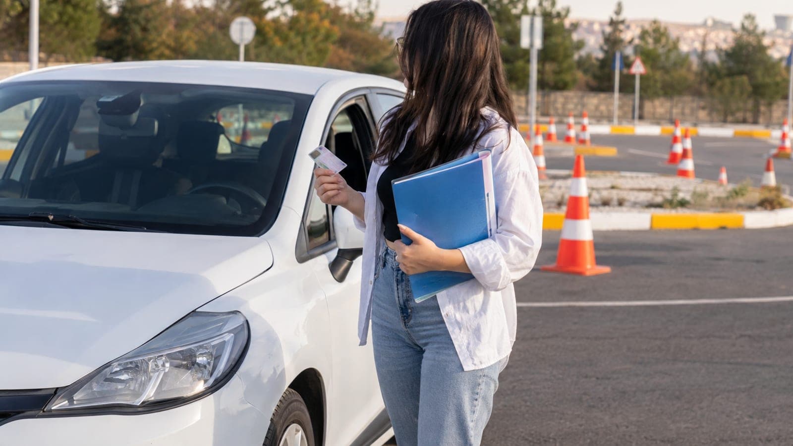 Sürücü kursu otoparkında eğitim otomobilinin yanında belge dosyası tutan genç bir kadın, arkada eğitim pisti konileri.
