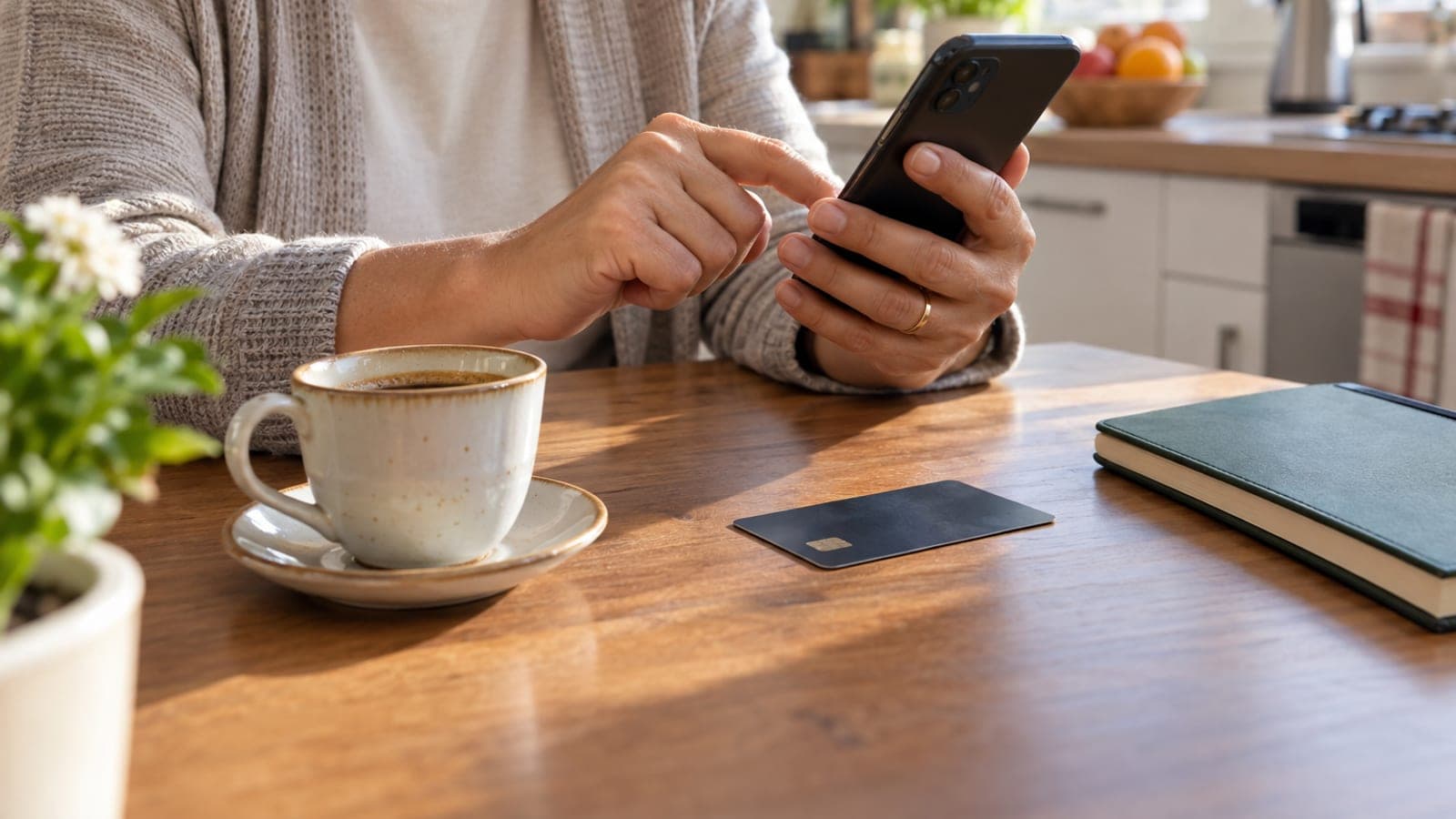 Mutfak masasında telefonuna dokunan bir kişi mobil bankacılık doğrulama ekranını kullanıyor; yanında banka kartı ve kahve fincanı var.