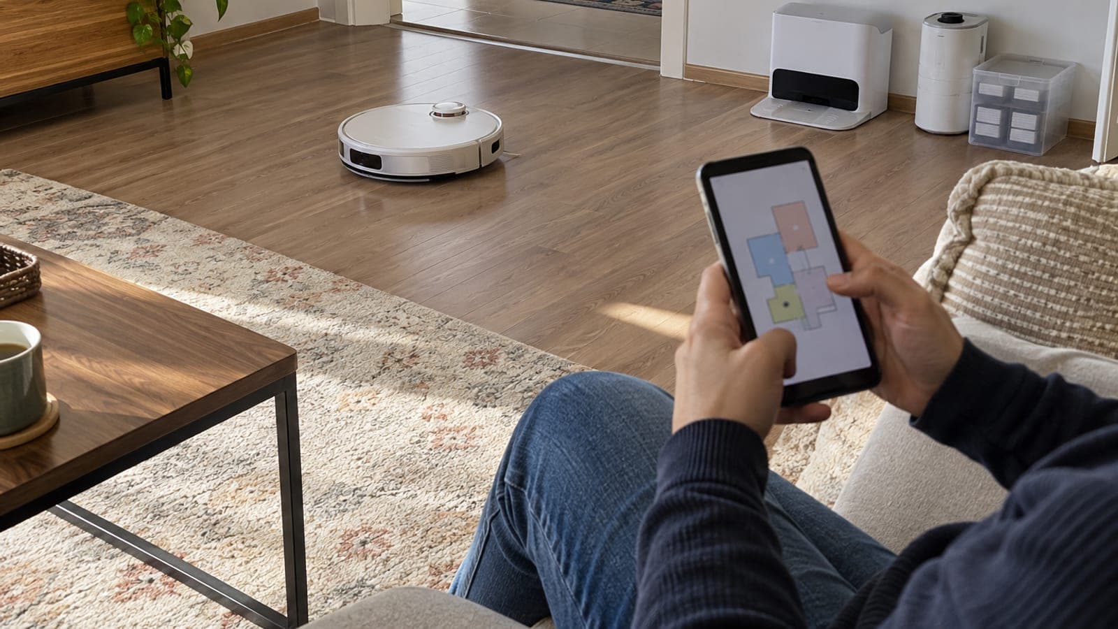 Laminat zeminde çalışan beyaz robot süpürge, duvardaki şarj istasyonu ve elinde telefonla uygulamayı kullanan kişi.