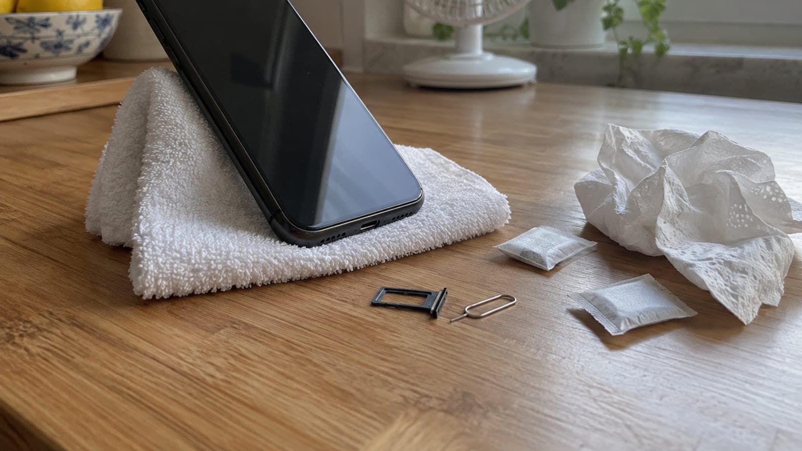 Mutfak tezgâhında havlu üstüne şarj girişi aşağı bakacak şekilde dayanmış telefon, yanında SIM tepsisi, iğne ve silika jel paketleri